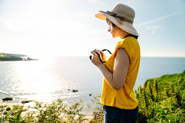 Случайно одетый блондин-турист в яркой желтой футболке и шляпе, захватывающий живописный прибрежный пейзаж с пышной зеленой скалы с полуденным светом - Фото, изображение
