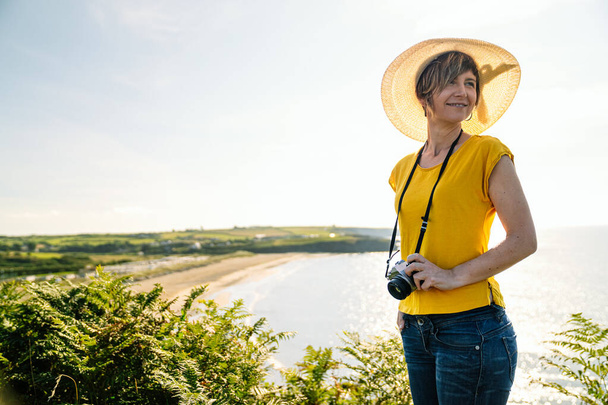 Eine lässige, lächelnde blonde Touristin in farbenfrohem gelben T-Shirt und Hut, mit der Kamera um den Hals, fängt die malerische Küstenlandschaft von einer saftig grünen Klippe unter der Mittagssonne ein.. - Foto, Bild