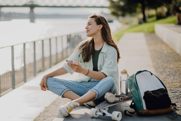 Onnellinen nuori nainen istuu kaupungin puistossa ja vierittää älypuhelinta. Nuorisokulttuuri ja työmatkaliikenne. - Valokuva, kuva
