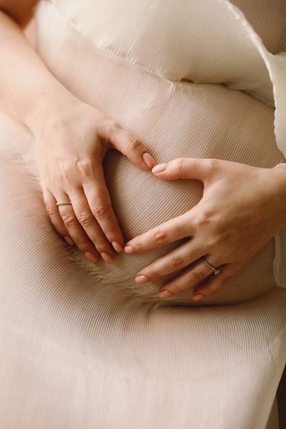 Zamknij zdjęcie kobiety w ciąży, która sprawia, że kształt serca z rąk na brzuchu. Kobieta oczekująca noworodka. Koncepcja macierzyństwa - Zdjęcie, obraz
