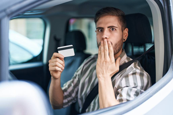 Hispanischer Mann mit Bart, der Auto fährt und Kreditkarten hält, die den Mund mit der Hand bedecken, schockiert und ängstlich vor Fehlern. Überraschte Miene  - Foto, Bild