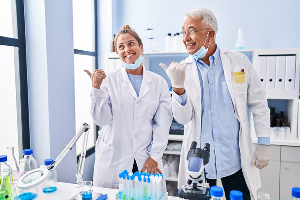 Middle age hispanic people working at scientist laboratory pointing thumb up to the side smiling happy with open mouth  - Foto, Bild