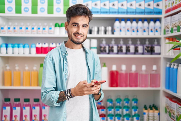 Nuori latino mies asiakas hymyilee luottavainen käyttämällä älypuhelin apteekissa - Valokuva, kuva
