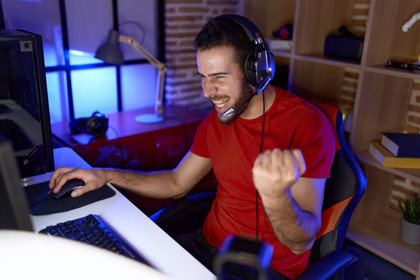 Jeune homme hispanique streamer jouer à un jeu vidéo avec l'expression gagnante à la salle de jeux - Photo, image