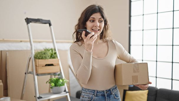 Mujer hispana hermosa joven que envía mensaje de voz por teléfono inteligente que sostiene el paquete en un nuevo hogar - Foto, Imagen
