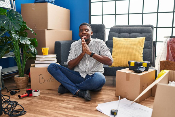 Αφροαμερικάνος κάθεται στο πάτωμα στο νέο σπίτι ικετεύοντας και προσευχόμενος με τα χέρια μαζί με την έκφραση της ελπίδας στο πρόσωπο πολύ συναισθηματική και ανησυχούν. επαιτεία.  - Φωτογραφία, εικόνα