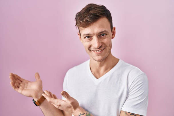Caucasian man standing over pink background inviting to enter smiling natural with open hand  - Photo, Image