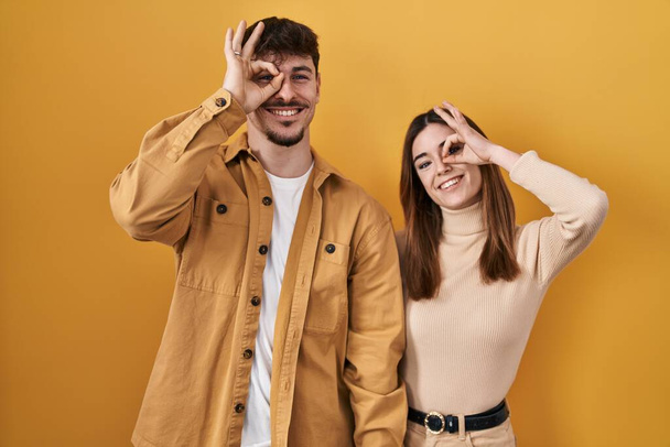 Junges hispanisches Paar, das über gelbem Hintergrund steht und mit lächelnder Hand ok-Geste macht, mit glücklichem Gesicht durch die Finger blickt.  - Foto, Bild