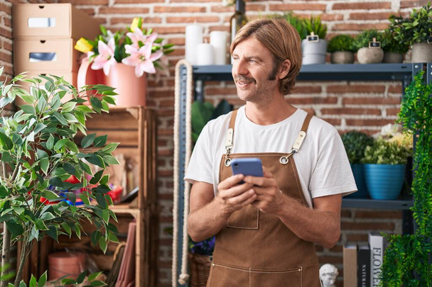 Nuori blondi mies kukkakauppias hymyilee luottavainen käyttäen älypuhelin kukkakauppa - Valokuva, kuva