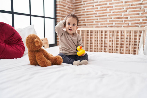 Schattige latino jongen met eend speelgoed zitten op bed in de slaapkamer - Foto, afbeelding