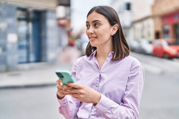 Junge hispanische Frau lächelt selbstbewusst mit Smartphone auf der Straße - Foto, Bild