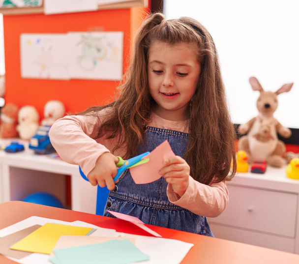 Adorable hispanic girl student smiling confident cutting paper at kindergarten - Photo, Image