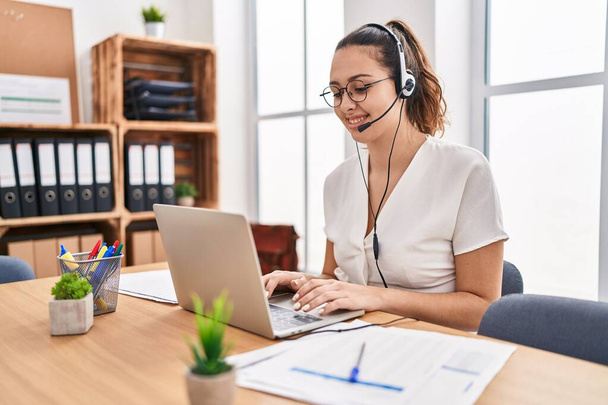 Mulher hispânica jovem vestindo fone de ouvido agente de call center no escritório olhando positivo e feliz de pé e sorrindo com um sorriso confiante mostrando dentes  - Foto, Imagem