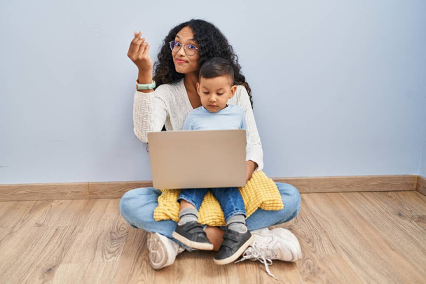 Jeune hispanique mère et enfant en utilisant ordinateur portable assis sur le sol faisant geste italien avec la main et les doigts expression confiante  - Photo, image