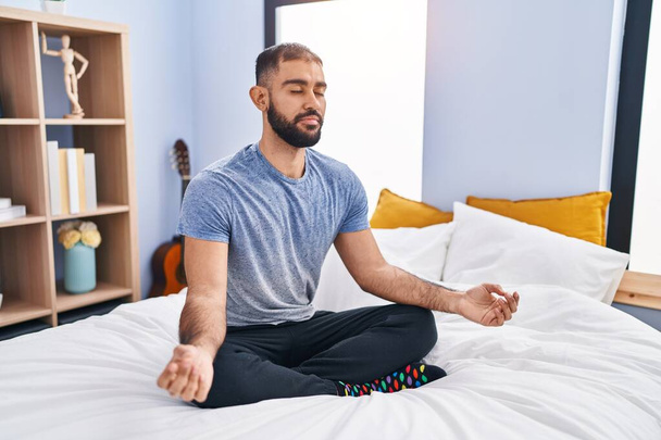 Young hispanic man doing yoga exercise sitting on bed at bedroom - Φωτογραφία, εικόνα