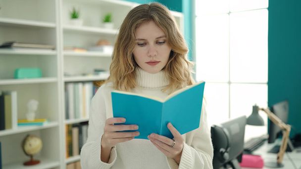 Young blonde woman student reading book standing at library university - Foto, imagen
