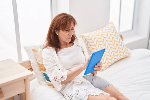 Μέση ηλικία γυναίκα χρησιμοποιώντας touchpad κάθεται στο κρεβάτι στο υπνοδωμάτιο - Φωτογραφία, εικόνα