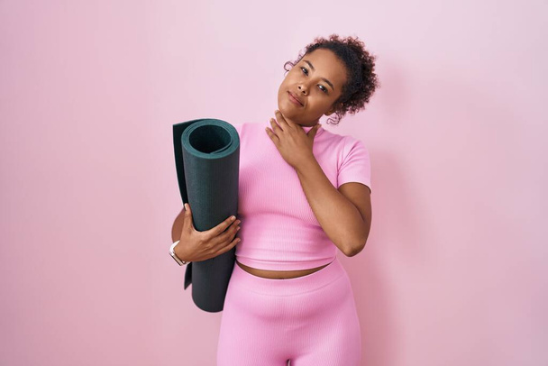 Junge hispanische Frau mit lockigem Haar hält Yogamatte über rosa Hintergrund und berührt schmerzhaften Hals, Halsschmerzen wegen Grippe, Scholle und Infektion  - Foto, Bild