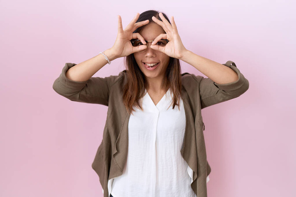 Middle age chinese woman wearing glasses over pink background doing ok gesture like binoculars sticking tongue out, eyes looking through fingers. crazy expression.  - Фото, зображення