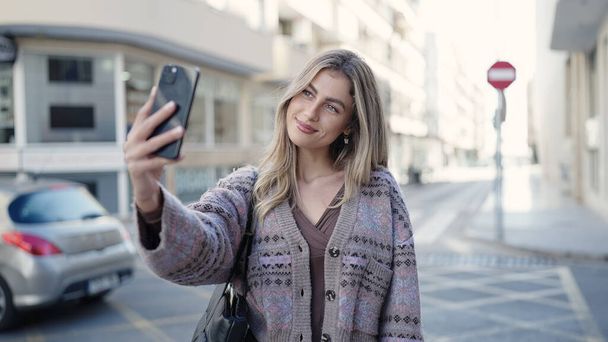 Genç sarışın kadın, kendinden emin gülümsüyor. Sokakta akıllı telefonun yanında özçekim yapıyor. - Fotoğraf, Görsel