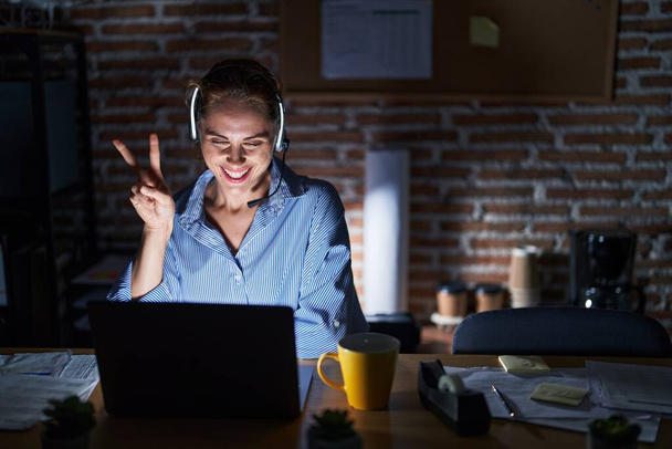 Beautiful brunette woman working at the office at night smiling with happy face winking at the camera doing victory sign. number two.  - Photo, Image