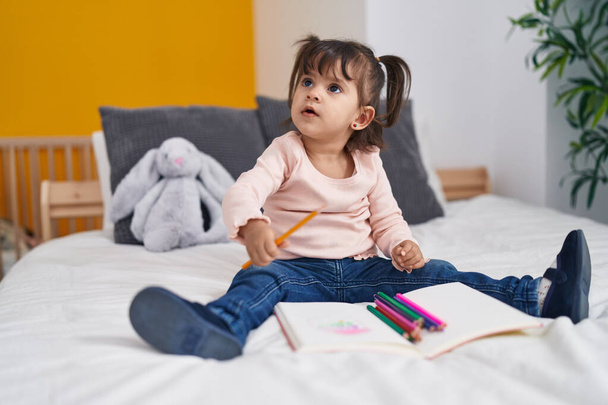 Adorable hispanic girl drawing on notebook sitting on bed at bedroom - Photo, Image