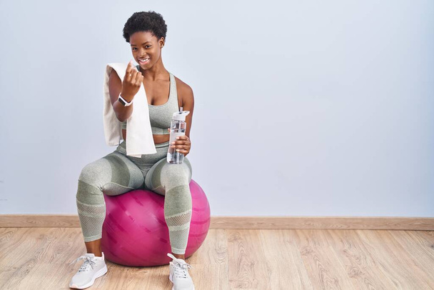 afro-americano mulher vestindo sportswear sentado no pilates bola acenando vir aqui gesto com a mão convidando recebimento feliz e sorrindo  - Foto, Imagem