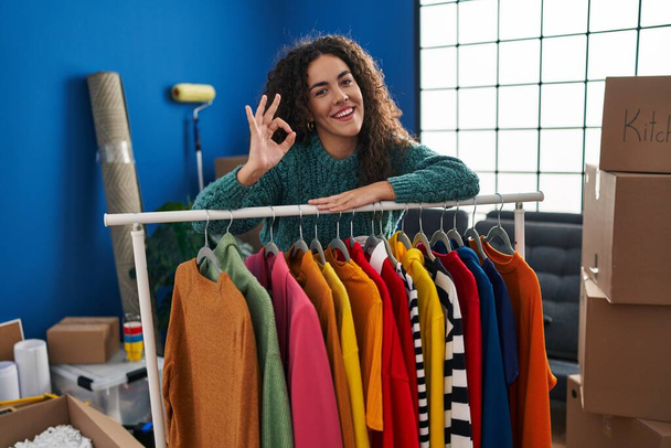 Nuori latino nainen seisoo uudessa kodissa vaatteet tekee ok merkki sormin, hymyillen ystävällinen ele erinomainen symboli  - Valokuva, kuva