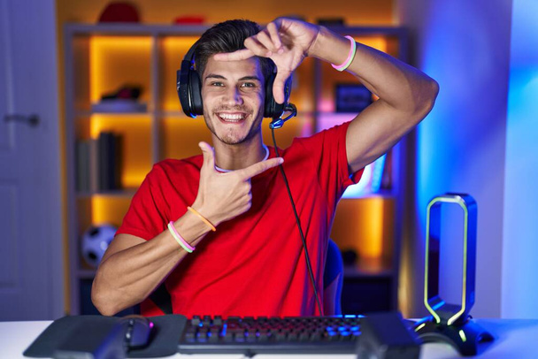 Junger hispanischer Mann, der Videospiele spielt, lächelnd Rahmen mit Händen und Fingern mit glücklichem Gesicht macht. Kreativität und Fotografiekonzept.  - Foto, Bild
