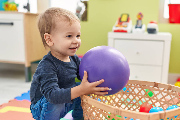 Imádnivaló szőke kisbaba, labdákkal játszik az óvoda padlóján ülve. - Fotó, kép