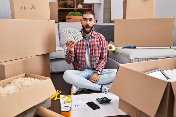 Hombre de Oriente Medio con barba sentado en el suelo en casa nueva sosteniendo el dinero asustado y sorprendido con la boca abierta para sorpresa, cara de incredulidad  - Foto, imagen