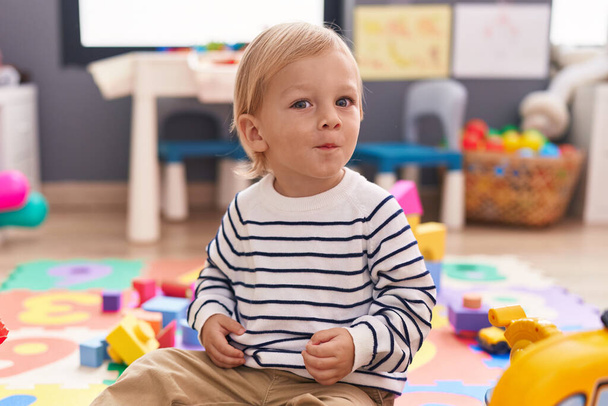 Adorable caucasian boy sitting on floor with relaxed expression at kindergarten - Foto, Imagem