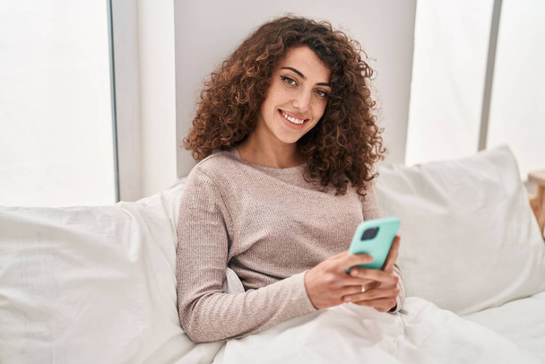 Молода іспаномовна жінка використовує смартфон, сидячи на ліжку в спальні
 - Фото, зображення