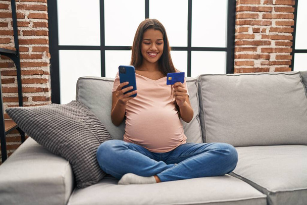 Young latin woman pregnant using smartphone and credit card at home - Foto, Bild