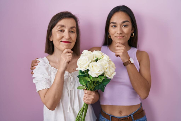 Mère et fille hispaniques tenant un bouquet de fleurs blanches regardant avec confiance la caméra souriant avec les bras croisés et la main levée sur le menton. penser positivement.  - Photo, image