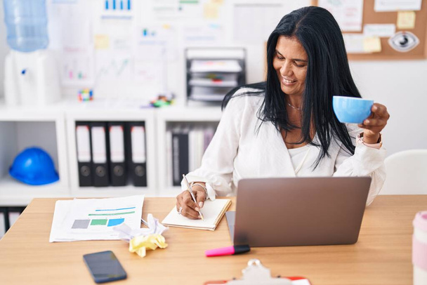 Middle age hispanic woman business worker writing on notebook drinking coffee at office - Photo, Image
