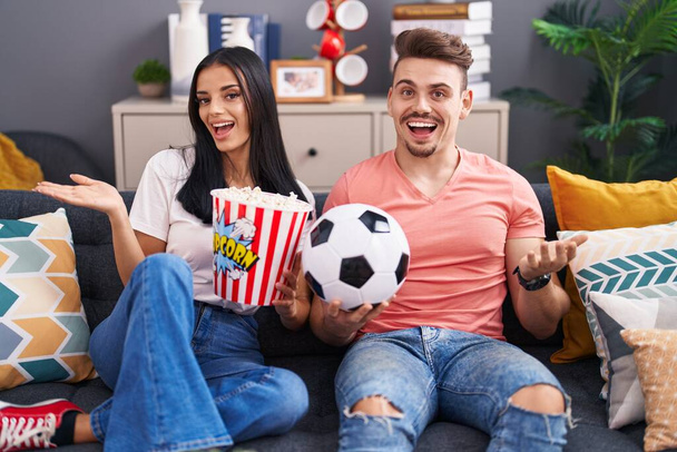 Fiatal spanyol pár futball huligánok gazdaság labda és eszik popcorn ünneplő teljesítmény boldog mosollyal és a győztes kifejezés emelt kézzel  - Fotó, kép