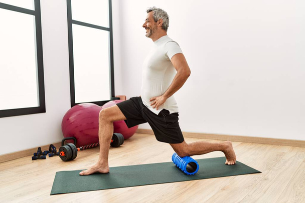Middle age grey-haired man using foam roller stretching at sport center - Foto, imagen