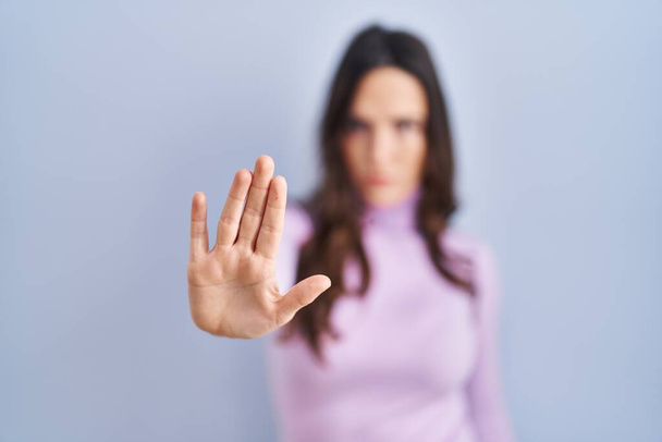 Young brunette woman standing over blue background doing stop sing with palm of the hand. warning expression with negative and serious gesture on the face.  - Foto, Imagen
