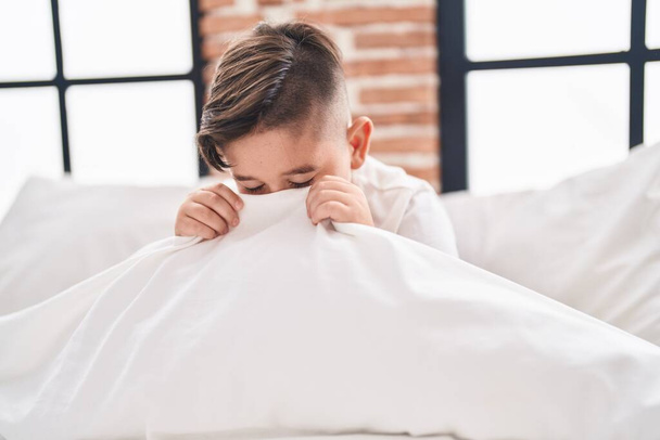 Adorable garçon hispanique assis sur le visage de couverture de lit avec drap de lit dans la chambre - Photo, image