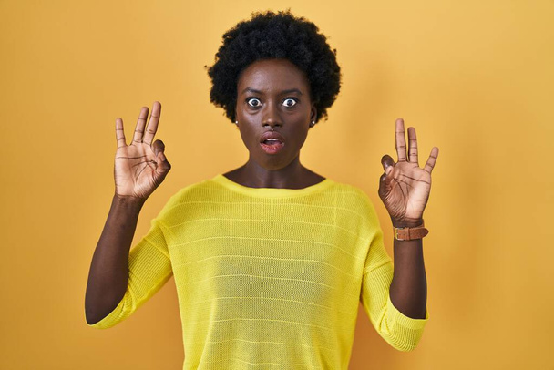 Jeune femme africaine debout sur studio jaune regardant surpris et choqué faire ok symbole d'approbation avec les doigts. expression folle  - Photo, image