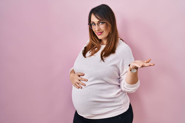 Zwangere vrouw staan over roze achtergrond glimlachend vrolijke presenteren en wijzen met palm van de hand kijken naar de camera.  - Foto, afbeelding