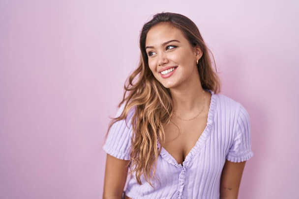 Junge hispanische Frau, die vor rosa Hintergrund steht und mit einem Lächeln im Gesicht zur Seite schaut, natürlicher Ausdruck. Lachen selbstbewusst.  - Foto, Bild