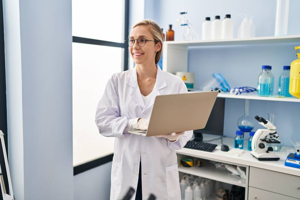 Junge blonde Wissenschaftlerin lächelt im Labor selbstbewusst mit Laptop - Foto, Bild