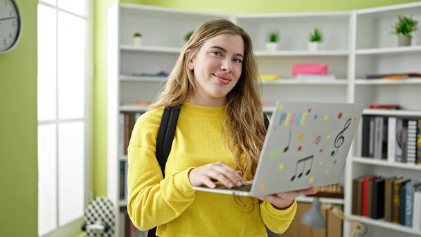 Jonge blonde vrouw student dragen rugzak met behulp van laptop op bibliotheek universiteit - Foto, afbeelding