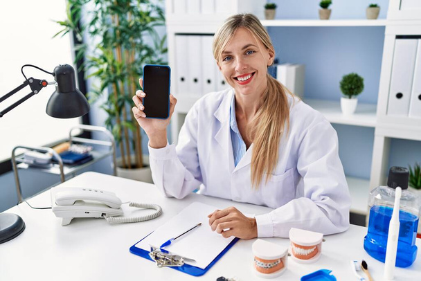 Kaunis hammaslääkäri nainen näyttää älypuhelin näyttö näyttää positiiviselta ja onnellinen seisoo ja hymyilee luottavainen hymy näyttää hampaat  - Valokuva, kuva