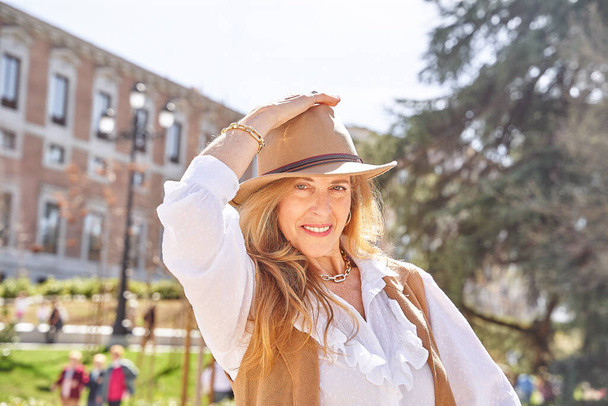  A carefree blonde woman with long hair and a stylish hat gazes directly into the camera, enjoying the peaceful surroundings of a park. - Photo, Image