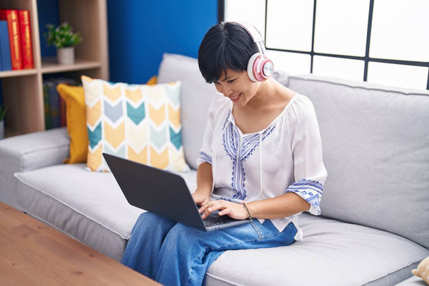 Donna cinese di mezza età che utilizza laptop e cuffie seduti sul divano a casa - Foto, immagini