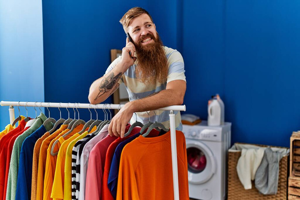 Mladý zrzek muž usmívá sebevědomě mluvit na smartphone v prádelně - Fotografie, Obrázek