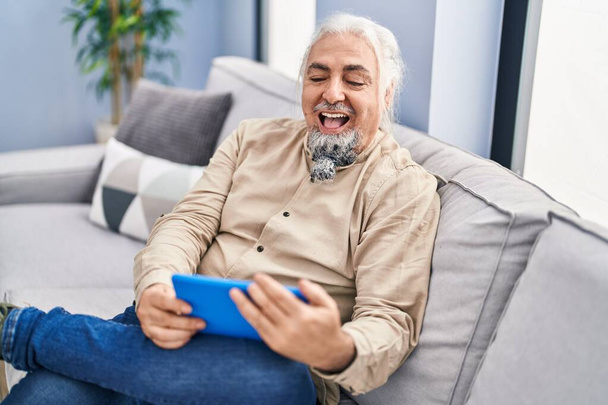 Middle age grey-haired man watching video on touchpad sitting on sofa at home - Photo, image